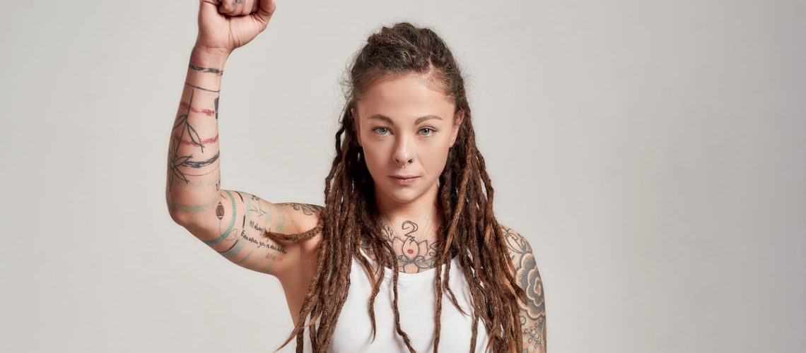 Portrait of young tattooed caucasian woman with dreadlocks wearing white shirt, looking confident at camera, raising her arm while posing isolated over grey background. Front view. Horizontal shot